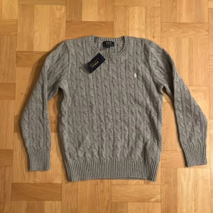 Grå kabelstickad tröja från Ralph Lauren - Säljer en stilren grå stickad tröja från Ralph Lauren med kabelstickat mönster. Tröjan är aldrig använd och har lappen kvar! 