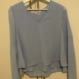 Ljusblå blus  - Säljer en elegant ljusblå blus från KappAhl med v-ringning och långa ärmar. Blusen har en fin volangdetalj vid nederkanten som ger en extra touch. Perfekt för både vardag och speciella tillfällen.