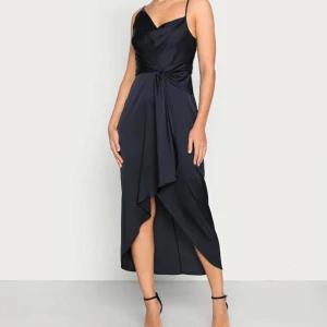 Satinklänning i mörkblått  - Elegant mörkblå klänning med smala asymmetriska axelband och v-ringning. Den har en stilren detalj framtill och en subtil slits som ger ett fint fall. Perfekt för en kväll ute eller en speciell tillställning. Axelbanden är justerbara. Nypris 1 300 kr. 