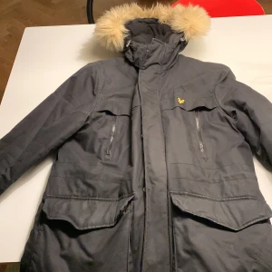 Svart parkajacka med pälsdetalj - Snygg svart parkajacka från Lyle & Scott med avtagbar huva och pälsdetalj. Jackan har dragkedja och knappar framtill samt flera praktiska fickor. Perfekt för kyliga dagar.