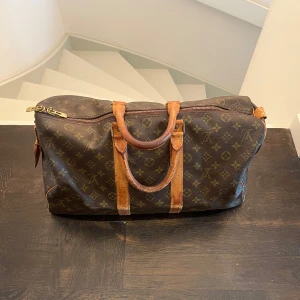  Louis Vuitton keepall size 50 - Säljer en klassisk brun monogramväska från Louis Vuitton med läderhandtag och detaljer. Väskan har en dragkedja upptill och är perfekt för resor eller som weekendbag. Den ikoniska designen gör den tidlös och stilren. ÄKTA GARANTERAT 
