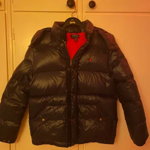  dunjacka från Polo Ralph Lauren - Säljer en marinblå dunjacka från Polo Ralph Lauren med röd insida. Jackan har en pufferdesign med långärmade ärmar och en praktisk huva. Den är utrustad med dragkedja och knappar för extra skydd mot kyla. Perfekt för kalla vinterdagar. Säljer nu den för att den inte passar mig och hoppas nån annan får användning av denna stilrena jacka i perfekt skick. Köpare står för frakten !