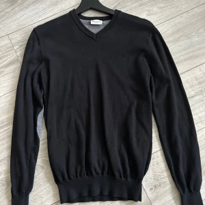 Svart tröja med grå armbågslappar från John Henric - Snygg svart tröja från John Henric med grå armbågslappar och v-ringad hals. Perfekt för en stilren look. Tröjan har långa ärmar och ribbade muddar vid ärmslut och nederkant.