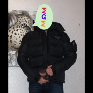 Marinblå pufferjacka Prada - Snygg marinblå pufferjacka med huva och dragkedja av märket Prada. Jackan har en quiltad design och en logotyp på bröstet. Perfekt för kyliga dagar.