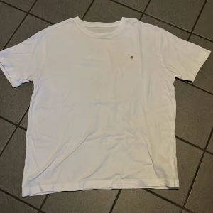 Vit t-shirt från GANT - Säljer en klassisk vit t-shirt från GANT med en liten logotyp på bröstet. Perfekt för en stilren och enkel look. T-shirten har korta ärmar och är tillverkad i mjukt bomullsmaterial.