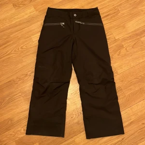 Svarta skid byxor från Marmot barn 128-134 - Säljer ett par svarta skid byxor från Marmot med dragkedjor framtill och baktill. Byxorna har en knapp i midjan och är perfekta för skidor eller utomhusaktiviteter. 