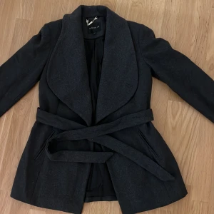 Svart kappa från Stockh LM - Elegant svart kappa från Stockh LM med ett stilrent bälte i midjan. Kappan har långa ärmar och en klassisk krage, perfekt för kyligare dagar. Den är tillverkad i ett mjukt material som ger en lyxig känsla.