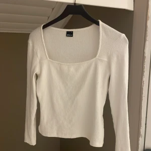 Vit ribbad topp från Gina Tricot - Säljer en stilren vit ribbad topp från Gina Tricot. Toppen har en fyrkantig halsringning och långa ärmar, perfekt för både vardag och fest. Den är i storlek M och passar utmärkt till olika outfits.