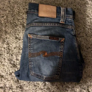 Blå jeans från Nudie Jeans - Säljer ett par snygga blå jeans från Nudie Jeans med klassisk femficksdesign och slitna detaljer. Jeansen har en slim ( grim tim ) passform och är tillverkade i denim med en autentisk tvätt för en cool look. Säljs för 399, nypris 1600.