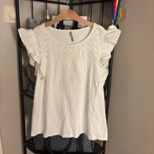 Vit blus med spetsdetaljer - Säljer en söt vit blus  med spetsdetaljer på ärmarna. Blusen har en rund halsringning och volangdetaljer som ger en romantisk touch. Perfekt för en somrig look!