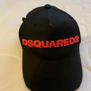 Svart keps från Dsquared2 - Snygg svart keps från Dsquared2 med röd broderad text framtill och baktill. Justerbar passform med spänne bak. Perfekt för en casual look. Bra skick PM vid frågor pris kan diskuteras