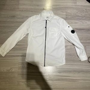 Vit overshirt från C.P. Company. - Säljer en stilren vit overshirt från C.P. Storlek 10 år men passar 140-160. Den sitter stort jag på bilden är 158 och kan ha den. Kvitto ingår. Köpt på nk rea för 1500 men nypris är 2500. Ser en liten fläck på bilden nu i efter hand men den borde gå att få bort i tvätt. Är inte helt säker men inget större att tänka på