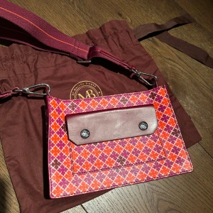 Färgglad axelväska By Marlene Birger - By Marlene Birger. Snygg axelväska med ett färgglatt rutigt mönster i rött, lila och orange. Väskan har en justerbar axelrem och en framficka med knappar. Använt 1 gång. 