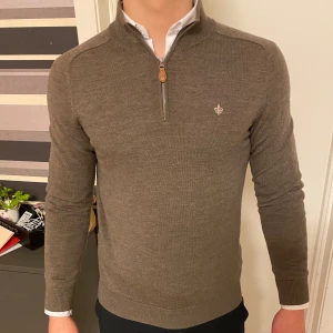 Brun tröja från Morris - Säljer en stilren brun tröja från Morris i 100% merinoull. Tröjan har en dragkedja vid halsen och ett diskret emblem på bröstet. Perfekt för kyligare dagar.