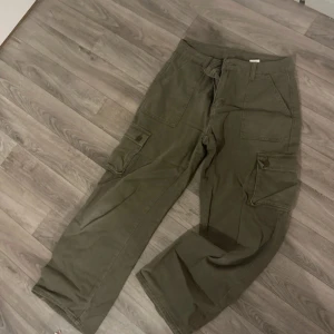 Olivgröna cargobyxor - Säljer ett par olivgröna cargobyxor med flera praktiska fickor och knappdetaljer. Byxorna har en loose passform och är perfekta för en avslappnad stil. De är tillverkade i ett slitstarkt material och har en midwaist men mer mot low waist på mig .