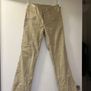Beige byxor med dragkedjedetaljer - Snygga beige byxor med raka ben och dragkedjor vid fickorna. Perfekta för en stilren look. De har en låg midja med stretchigt material.