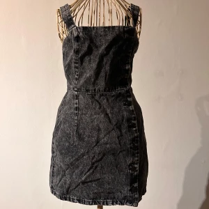 Svart jeansklänning från Divided - Trendig svart jeansklänning med knappar längs hela sidan. Klänningen har axelband och en figurnära passform som ger en snygg siluett. Perfekt för en cool och avslappnad stil.