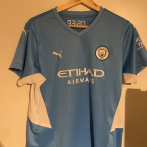 Ljusblå Manchester City fotbollströja  - Säljer en ljusblå Manchester City fotbollströja från Puma med korta ärmar. Tröjan har klubbens emblem på bröstet och sponsorlogga 'Etihad Airways' framtill. Perfekt för fans av laget! Liten i storlek 