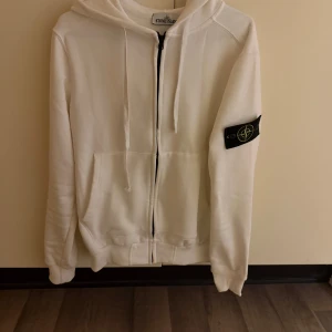 Vit hoodie från Stone Island - Säljer en snygg vit hoodie från Stone Island. Den har en dragkedja framtill och en klassisk känguruficka. Perfekt för en stilren och avslappnad look. Märkesloggan sitter på ärmen. Passar perfekt till både vardag och chill.