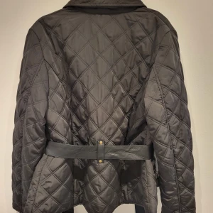 Svart quiltad jacka med bälte - Snygg svart quiltad jacka  med bälte i midjan och dragkedja framtill. Jackan har två bröstfickor med knappar och en cool camouflagemönstrad insida. Perfekt för en stilren look!