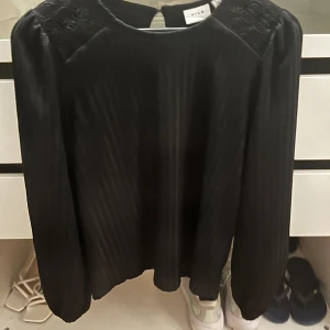 Svart blus från VILA - Elegant svart blus från VILA med långärmade puffärmar och subtila ribbade detaljer. Perfekt för en stilren look med en knappdetalj i nacken.