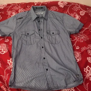 Randig skjorta från Jack & Jones - Säljer en stilren blå och vit randig skjorta från Jack & Jones. Skjortan har korta ärmar och två bröstfickor med knappar. Perfekt för en avslappnad look.