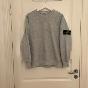 Grå sweatshirt från Stone Island - Säljer en stilren grå sweatshirt från Stone Island med deras ikoniska märke på ärmen. Tröjan har en klassisk rund hals och långa ärmar, perfekt för en avslappnad look. Knappt använd och i mycket bra skick. Storlek M, men passar även bra som S 