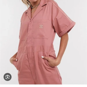 Rosa jumpsuit från Adidas - slutsåld överallt! Säljer en supercool rosa jumpsuit från Adidas. Den har korta ärmar och en avslappnad passform med praktiska fickor fram och bak.  Adidas-loggan är snyggt placerad på bröstet. 