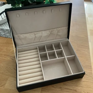 Svart smyckeskrin med fack - Säljer ett stilrent svart smyckeskrin med flera fack för smycken och klockor. Insidan är klädd i beige tyg och har plats för ringar och andra småsaker. Perfekt för att hålla ordning på dina accessoarer!