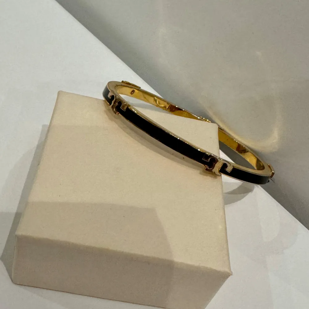 Använd bara ett fåtal gånger! Elegant svart och guld emaljerat armband med Tory Burch-logga. Ett mångsidigt tillbehör för alla garderober. • Svart och guld emalj • Tory Burch-logga • Hängslenstängning • Högkvalitativ finish • Lättviktsdesign. Asusteet.