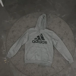 Grå hoodie från Adidas - Säljer en grå hoodie från Adidas med stor logga på framsidan. Tröjan har en klassisk känguruficka och justerbar huva. Perfekt för en avslappnad stil.