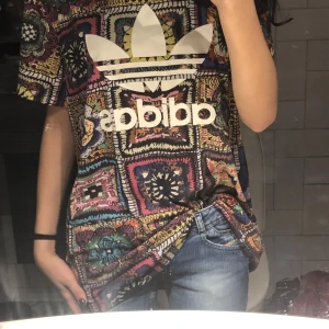 Mönstrad t-shirt från Adidas - Säljer en färgglad och mönstrad t-shirt från Adidas med stort logotryck på framsidan. T-shirten har korta ärmar och ett unikt, bohemiskt mönster i flera färger som lila, blå, gul och röd. Perfekt för dig som vill sticka ut!