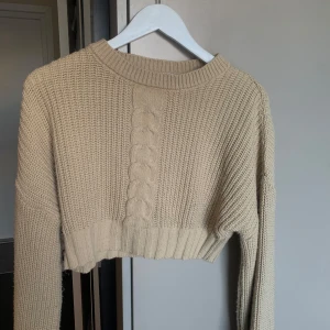 Beige stickad croppad tröja - Säljer en beige stickad tröja med ett snyggt flätmönster framtill. Tröjan är croppad och har långa ärmar, perfekt för en trendig look. Passar bra till både jeans och kjol!