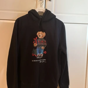 Svart hoodie från Polo Ralph Lauren med nallemotiv - Säljer en svart hoodie från Polo Ralph Lauren med ett gulligt nallemotiv på framsidan. Hoodien har en klassisk design med långa ärmar och en bekväm huva. Perfekt för en avslappnad stil. Tröjan är i storlek S och Andvänd ett fåtal gånger skicket är 9/10, nypris är runt 2700
