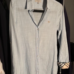 Ljusblå linneskjorta från Burberry - Snygg ljusblå skjorta i linne från Burberry med klassisk krage och knappar framtill. Skjortan har långa ärmar och ett diskret broderat emblem på bröstet. Perfekt för en stilren look. Det står att den är i storlek M fast den sitter som en XS eftersom den har krympt i tvätten. Hör av dig vid andra frågor😁😁