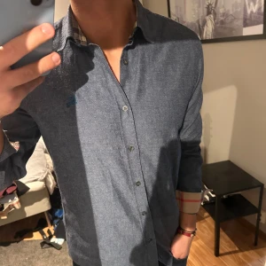 Burberry skjorta - Snygg blå skjorta från Burberry Brit med klassisk krage och knappar framtill. Skjortan har långa ärmar och ett diskret rutigt mönster på insidan av kragen och ärmsluten. Perfekt för en stilren look.
