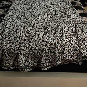 Blommig klänning med volangdetaljer - Charmig klänning med svart bas och vita blommor. Den har volangdetaljer och en v-ringad hals. Perfekt för en somrig look.