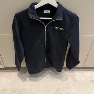 Svart jacka från Les Deux - Snygg svart jacka från Les Deux med dragkedja framtill. Jackan har en stilren design med långärmade ärmar och en liten logotyp på bröstet. Perfekt för en casual look.