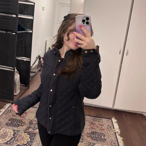 Svart quiltad jacka från Zara Woman - Snygg svart quiltad jacka från Zara Woman med knappar och hög krage. Jackan har långa ärmar och stilrena detaljer i brunt längs kanterna. Perfekt för kyligare dagar.