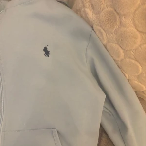 Ljusblå hoodie från Ralph Lauren - Säljer en stilren ljusblå hoodie från Ralph Lauren med dragkedja och det klassiska logotypbroderiet på bröstet. Perfekt för en avslappnad stil.