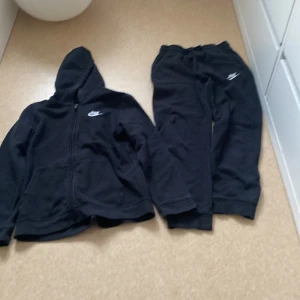 Svart hoodie och byxa från Nike - Säljer en svart hoodie från Nike med dragkedja och huva. Den har en broderad Nike-logga på bröstet och är tillverkad i ett mjukt material. Perfekt för en avslappnad stil.