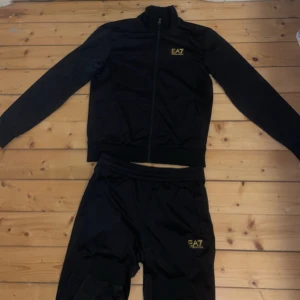 Svart träningsset från EA7 Emporio Armani - Snyggt svart träningsset från EA7 Emporio Armani med gulddetaljer. Setet består av en jacka med dragkedja och matchande byxor. Perfekt för en stilren look på gymmet eller till vardags.