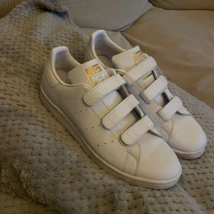 Vita Adidas Stan Smith med kardborreband - Säljer ett par klassiska vita Adidas Stan Smith sneakers med kardborreband. Skorna har en stilren design med guldiga detaljer på plösen och hälen. Perfekta för en avslappnad stil.