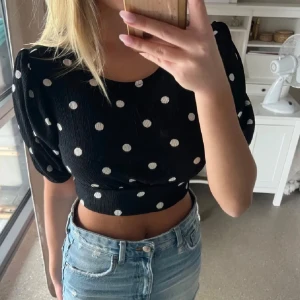 Svart prickig croppad topp - Snygg svart croppad topp med vita prickar och korta puffärmar. Toppen har en öppen rygg med knytband som ger en extra detalj. Perfekt för en trendig look!