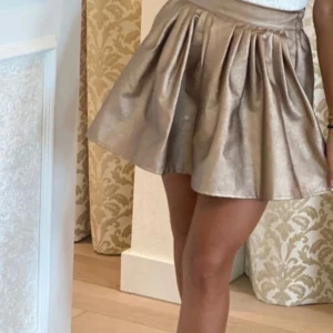 Guldglänsande kjol från Merry Kohn - Snygg guldglänsande kortkjol från Merry Kohn i storlek L. Kjolen har en elastisk midja för bekväm passform och är tillverkad i ett glansigt material av polyester och elastan. Perfekt för att ge en touch av glamour till din outfit!