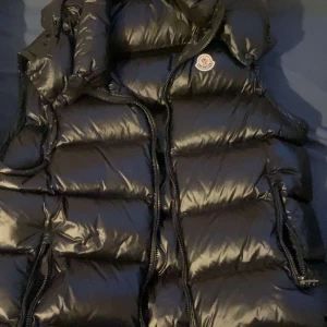 Svart dunväst från Moncler - Säljer en snygg svart dunväst från Moncler med glansig yta och dragkedja framtill. Västen har en hög krage och Moncler-logga på bröstet. Perfekt för kyliga dagar!