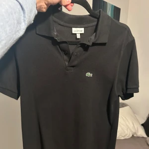 Svart pikétröja från Lacoste - Snygg svart pikétröja från Lacoste med klassisk krage och knappslå. Perfekt för en stilren look. Krokodillogga på bröstet. Passar både till vardag och mer uppklädda tillfällen. Liten i storlek. Mer bilder och beskrivning finns om så önskas. Pris kan diskuteras:) Mvh/ Philip