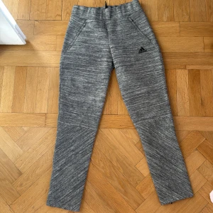 Grå träningsbyxor från Adidas - Snygga grå träningsbyxor från Adidas med dragkedja vid bensluten. Perfekta för träning med en bekväm passform och elastisk midja. Adidas-logga på framsidan ger en sportig touch.