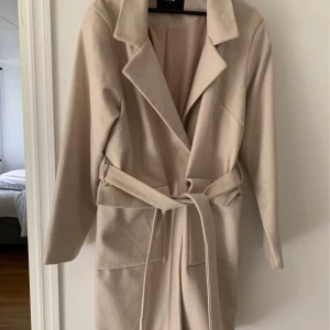 Beige kappa från Bik Bok - Elegant beige kappa från Bik Bok med knytband i midjan och långa ärmar. Perfekt för höst och vår, med en stilren design och praktiska fickor framtill.