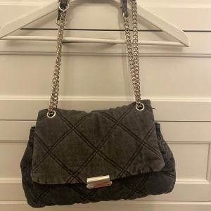 Svart axelväska med kedjedetaljer - Snygg svart-grå  quiltad väska. Man kan ha den på axeln och crossover. Tyget är denimaktigt. Rymlig, får plats med mycket grejer. Väldigt fint skick.